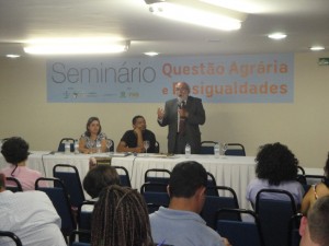 O ministro Gilberto Carvalho, falando ao microfone. À esquerda, sentado, com a mão no rosto, Pedro Stédile líder do MST